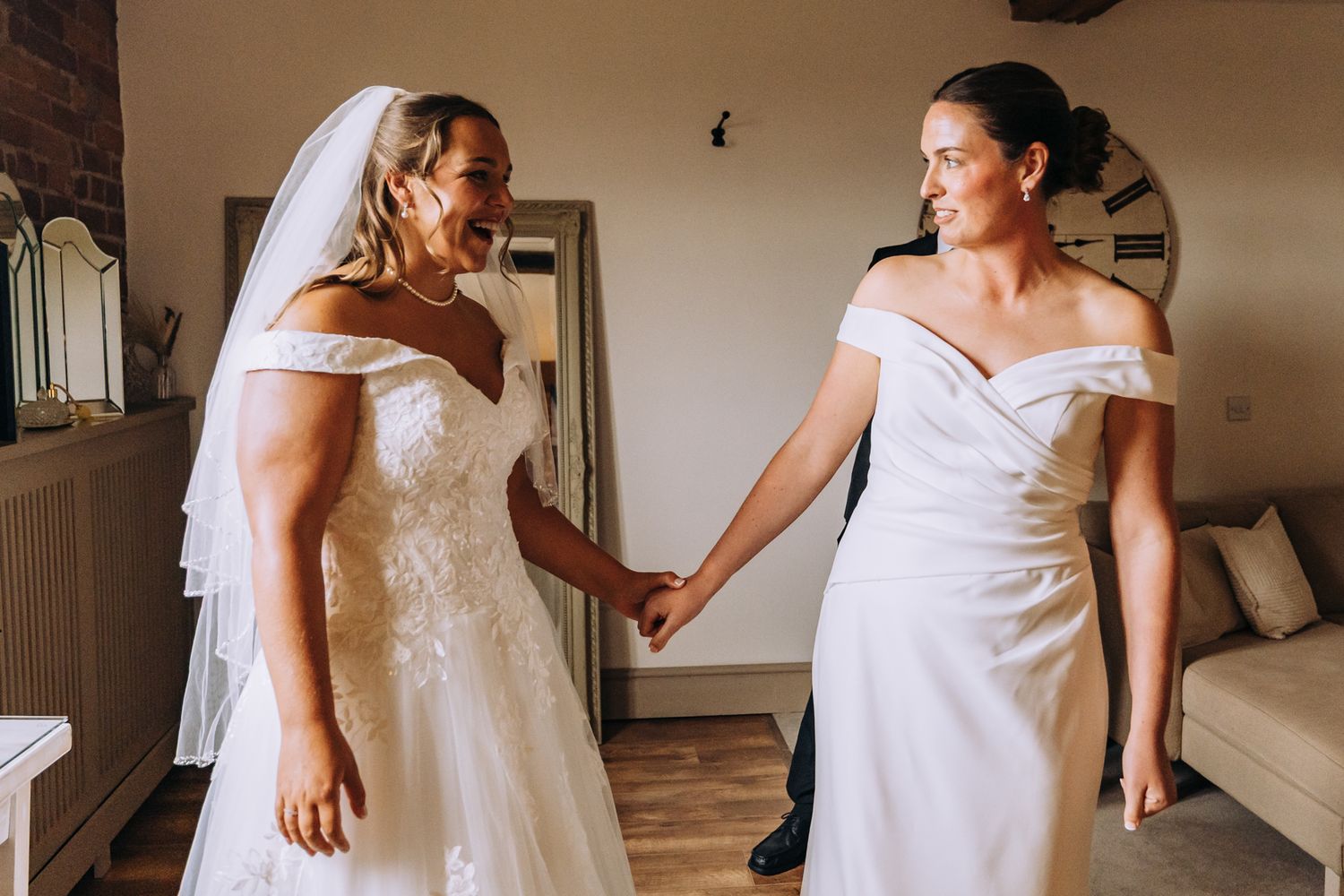 Two people in white dresses laugh together during wedding preparations.
