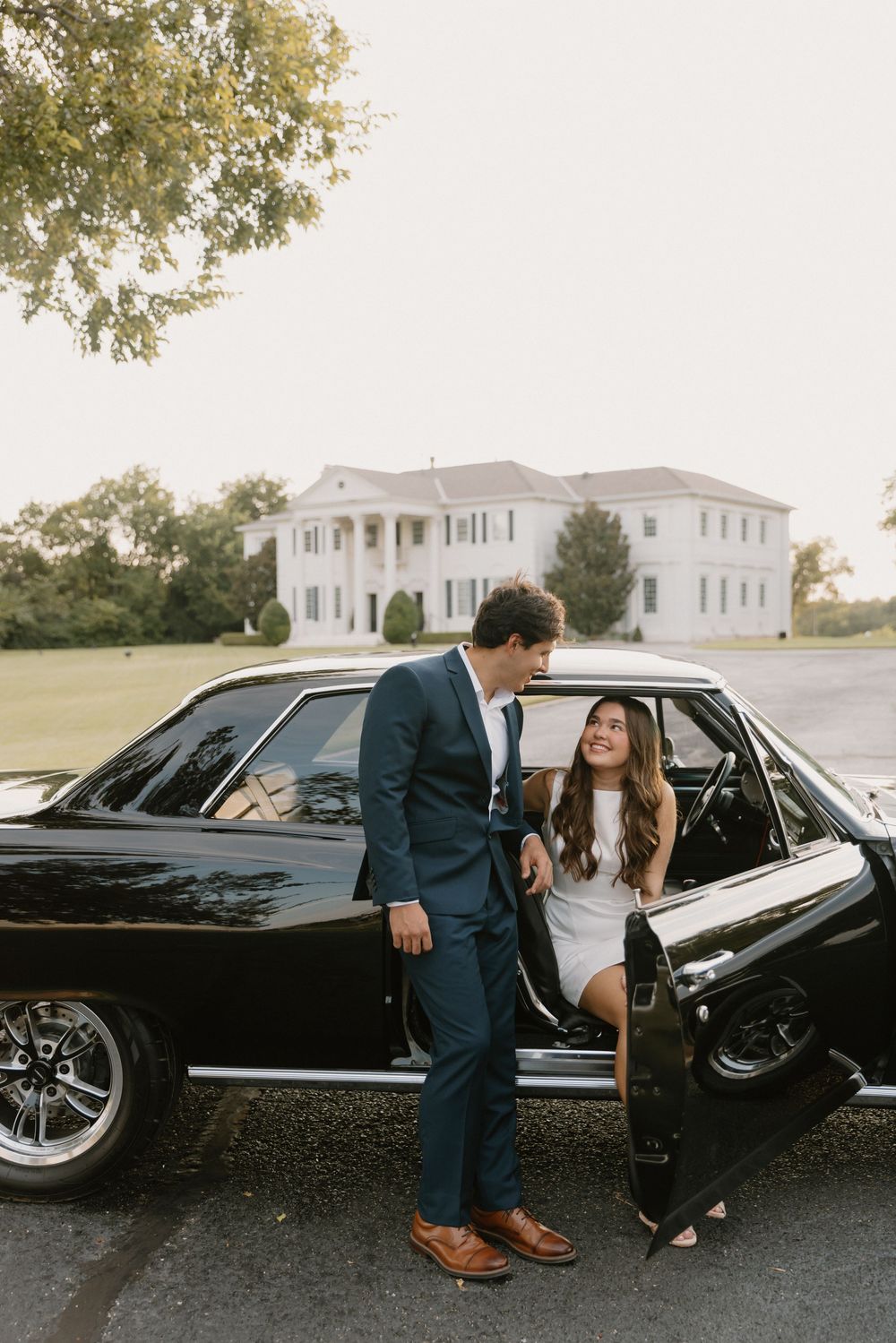 Romantic couple poses with vintage black car outside white mansion at sunset.