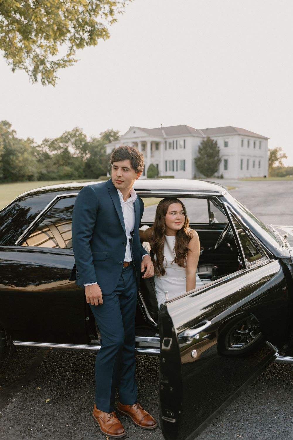 A couple poses next to a classic black car during a romantic photoshoot at sunset.
