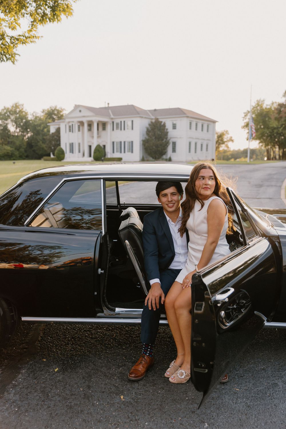 A couple sits together in a vintage black car parked outside a white mansion on a sunny day.