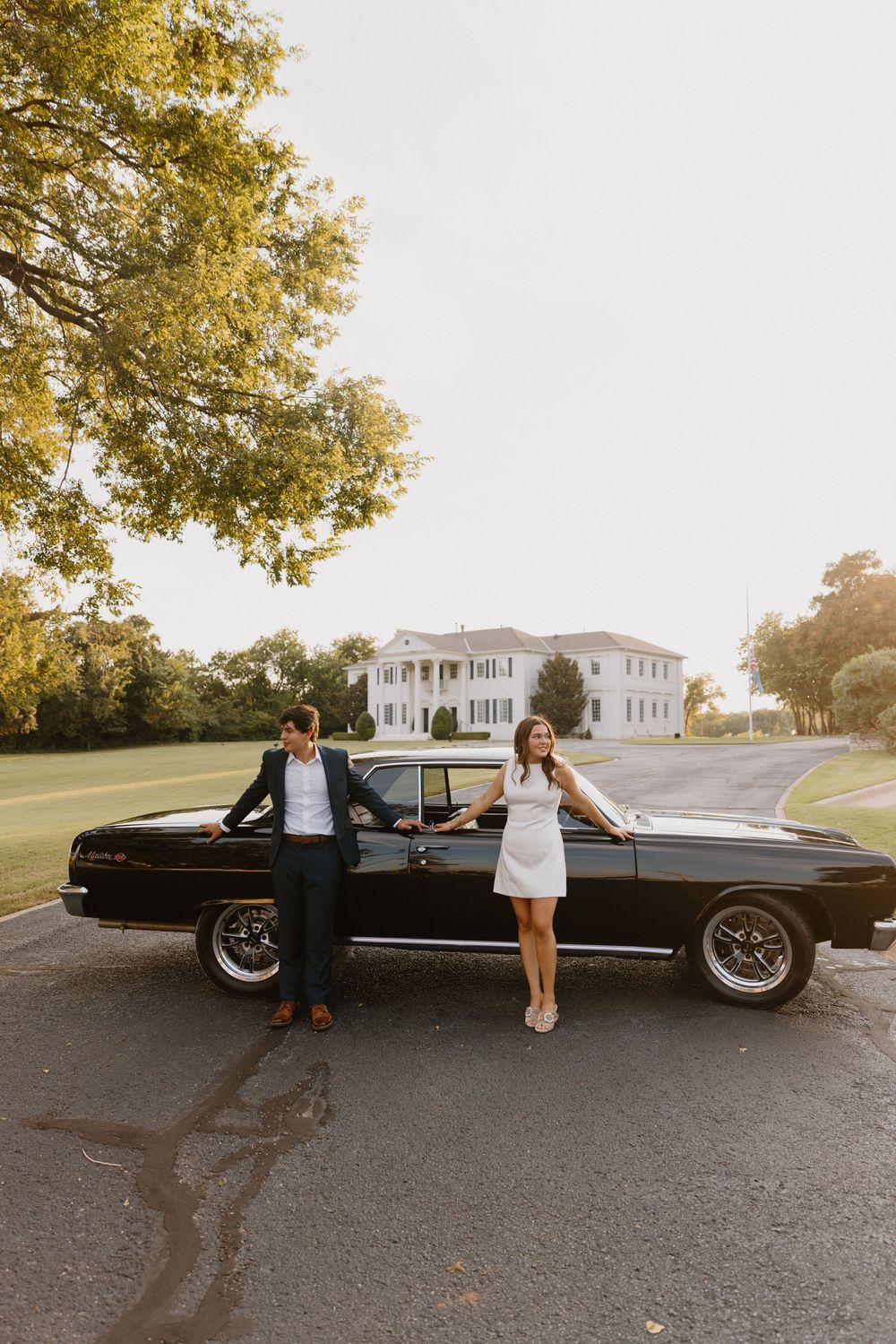 Classic black car parked in front of elegant white colonial mansion with couple posing.