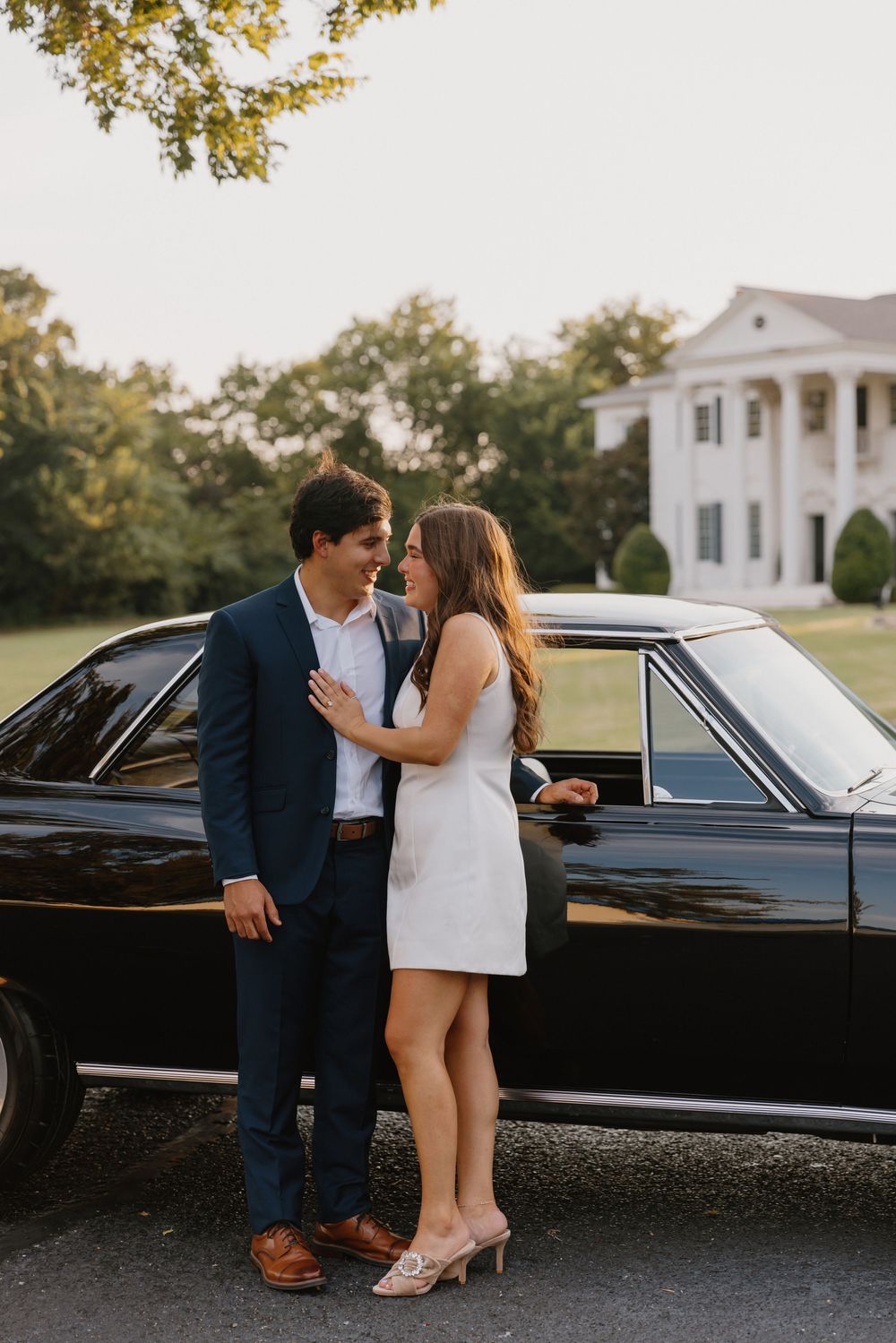 A romantic couple shares an intimate moment beside a vintage black car on a sunny afternoon.