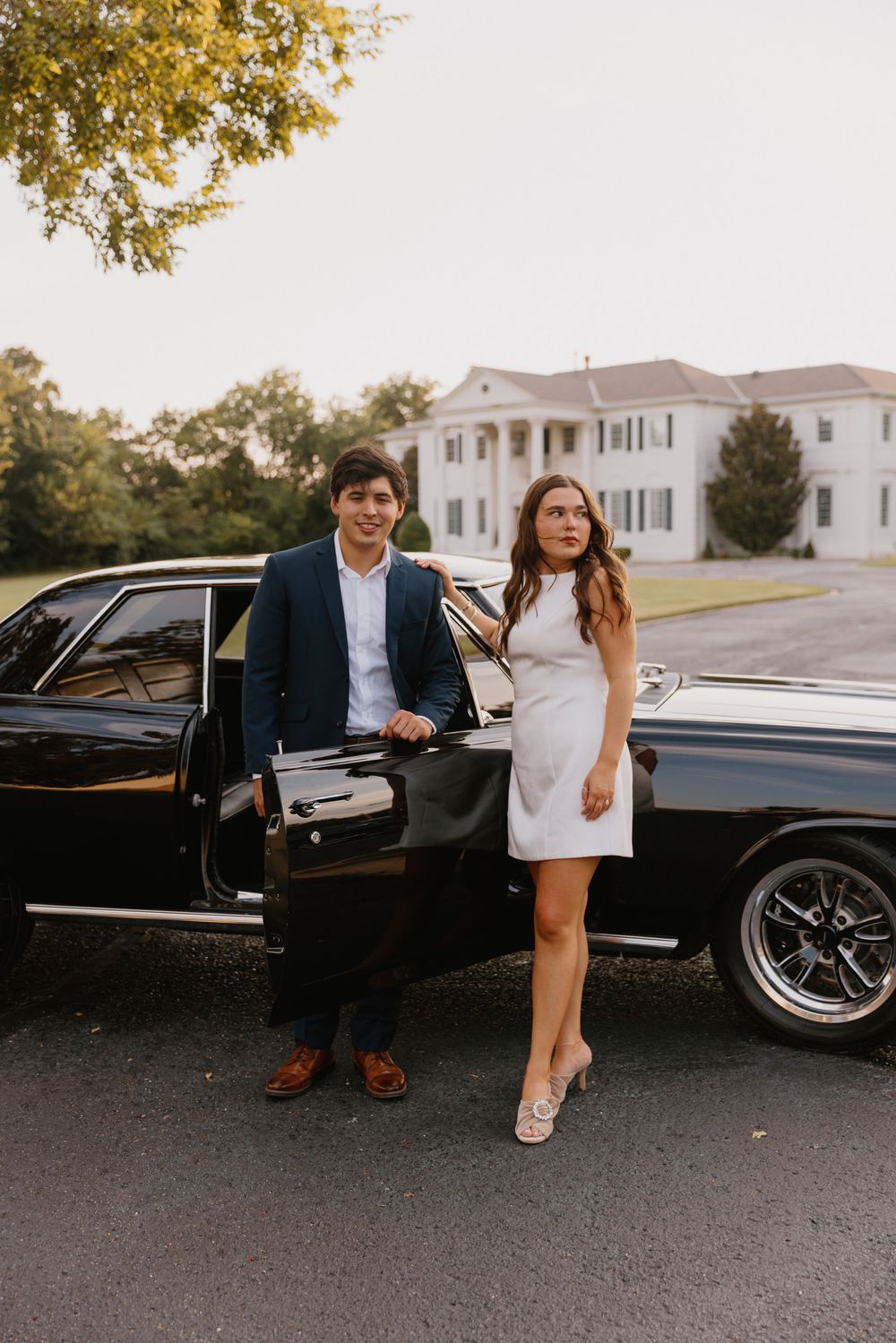 A couple poses elegantly next to a black luxury car on a paved driveway during a formal event.
