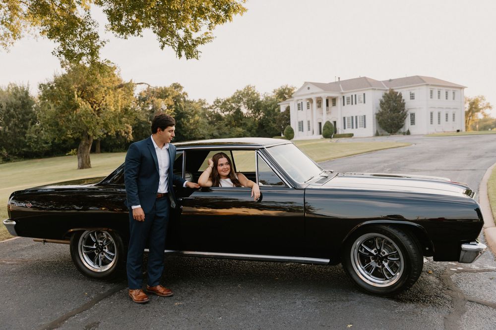 Classic black muscle car parked in front of a stately white mansion on a sunny autumn day.