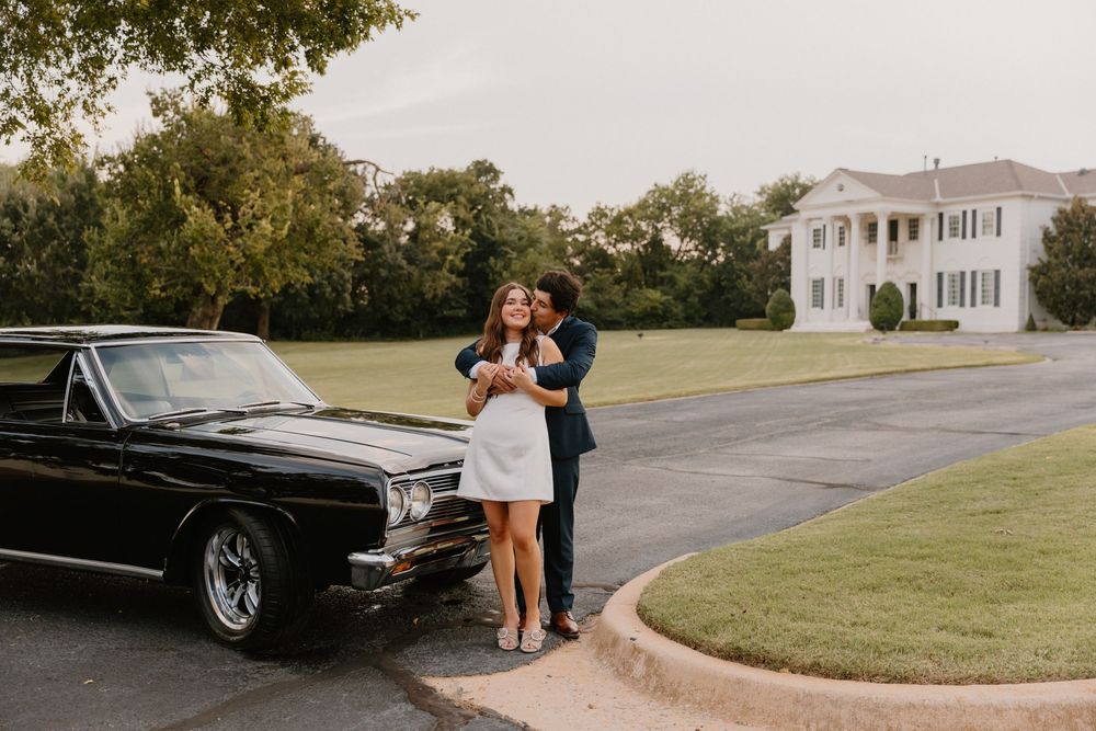 A black classic car is parked on a paved driveway near a grassy lawn.