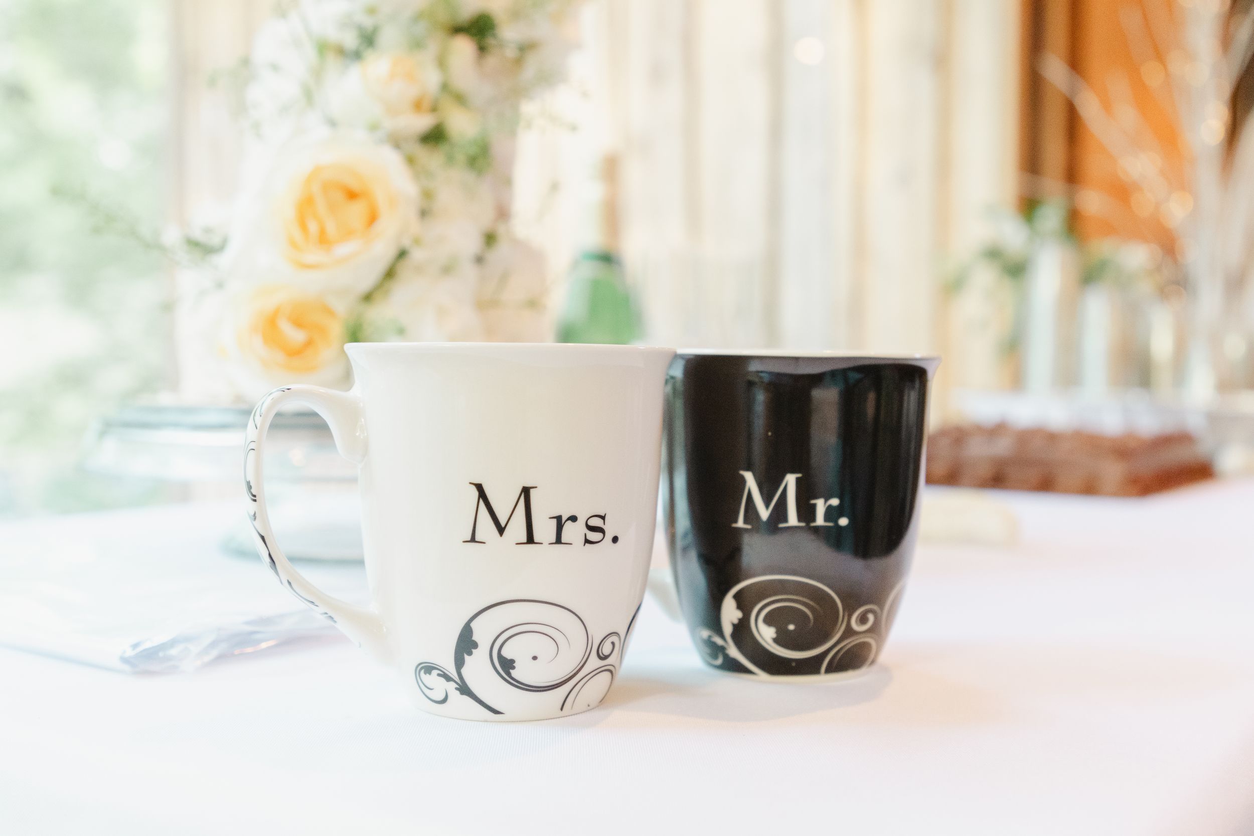 Black and white Mr. and Mrs. coffee mugs with decorative swirls displayed with yellow roses.