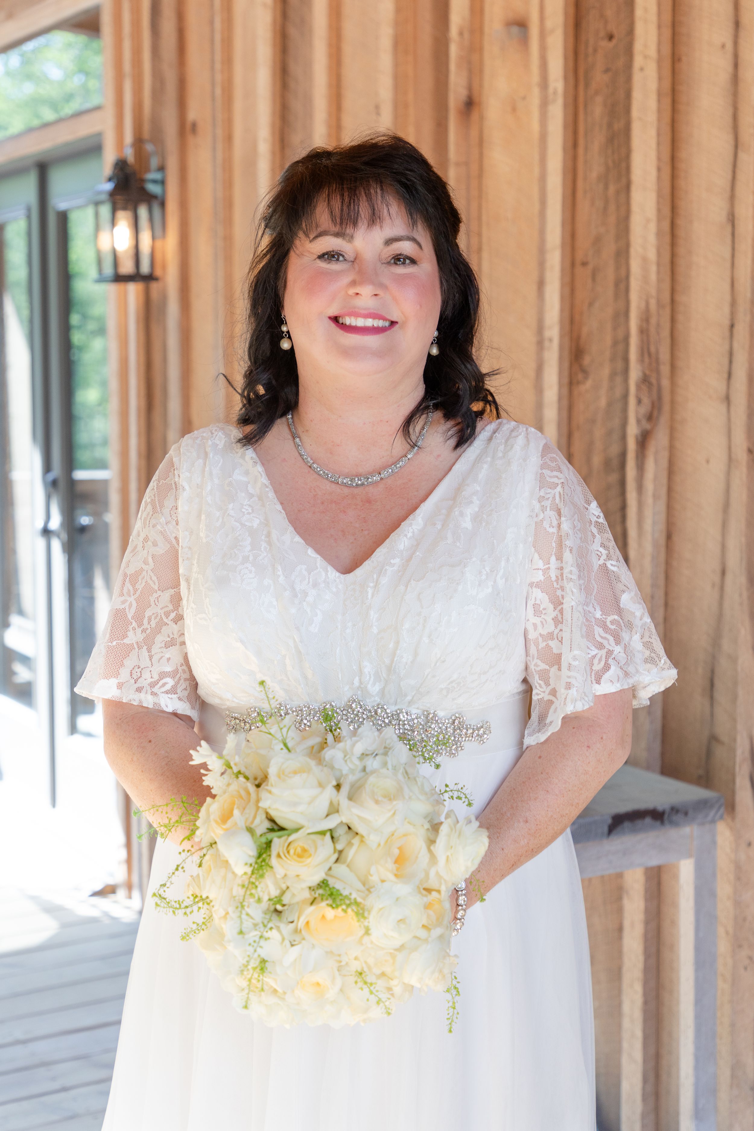 A bride in a lace-sleeved white wedding dress holds a bouquet of yellow and white roses.