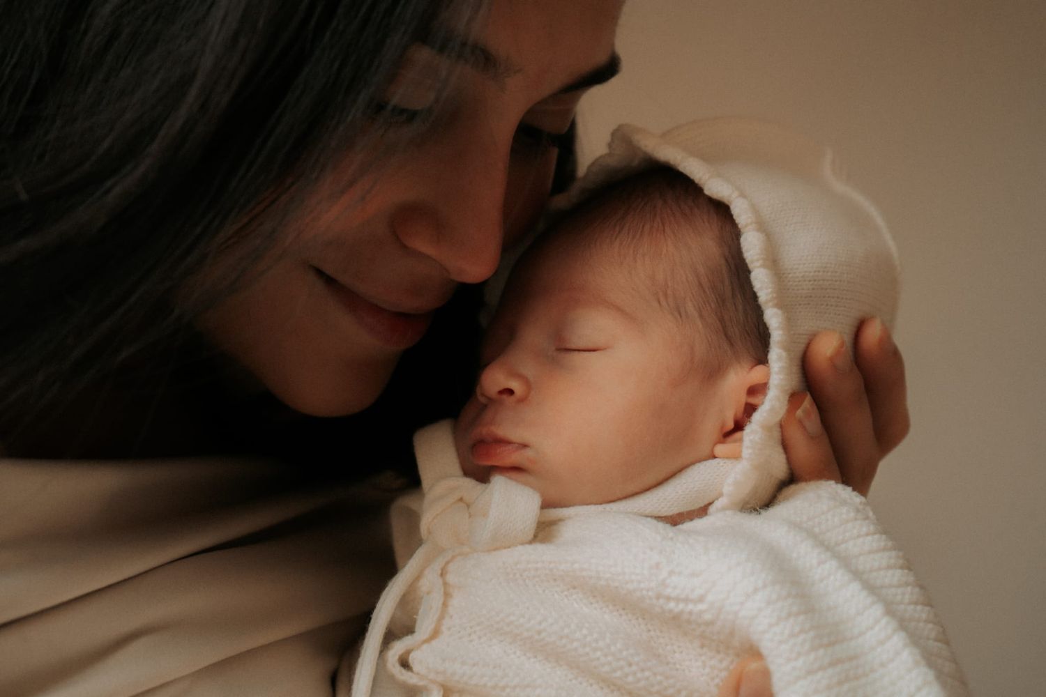 A mother gently nuzzles her peaceful sleeping newborn dressed in a white bonnet and swaddle.