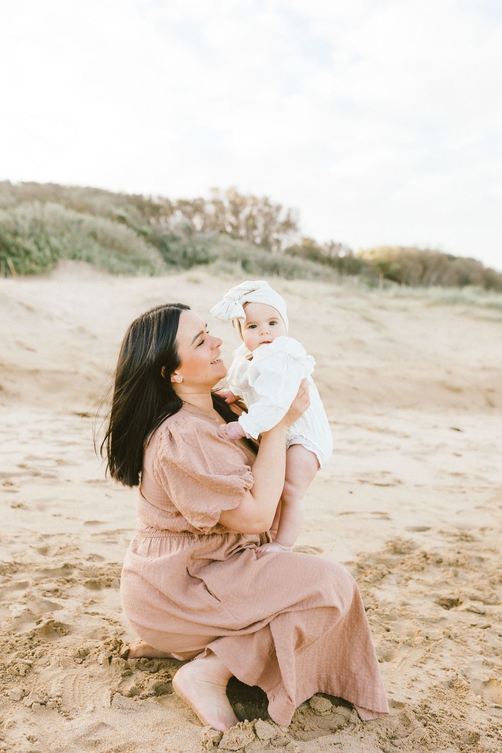 A parent in a pink dress plays lovingly with a baby on a sandy beach during a sunny day.