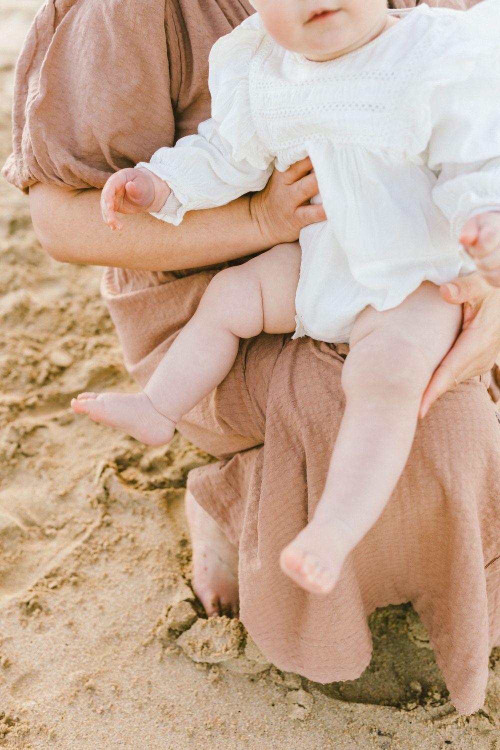 Close-up view of a person in brown linen pants sitting on sandy ground while holding a child in a white dress.