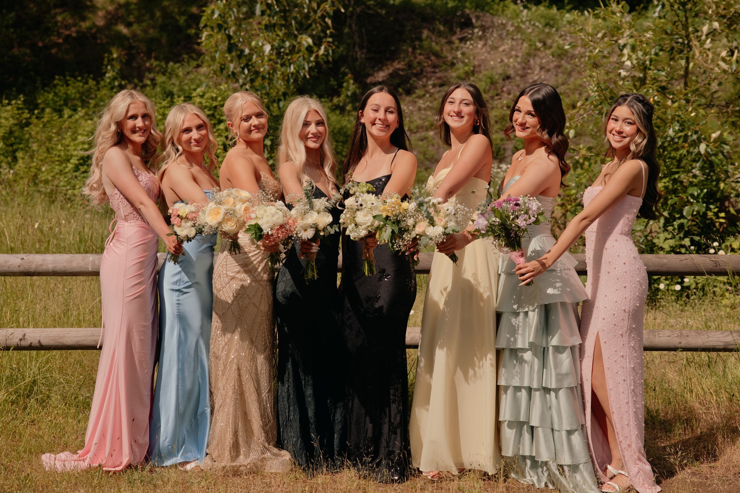 Seven bridesmaids in mismatched pastel gowns holding floral bouquets pose together outdoors near a wooden fence.