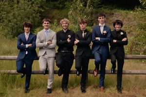Six young men in formal suits posing together leaning on a wooden fence outdoors.
