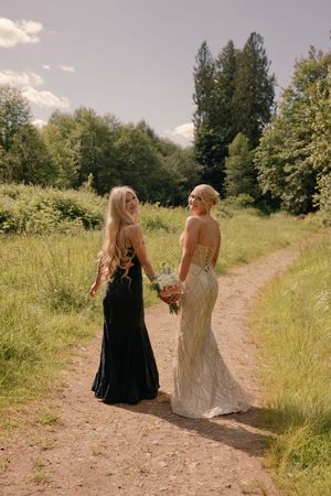 Two blonde women in elegant gowns, one in black and one in white bridal dress, walking a scenic garden path.