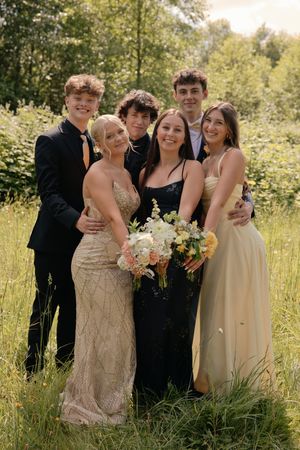 Six teens in formal attire pose together in a lush green field, smiling with bouquets at prom.