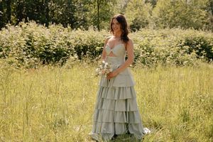 Young woman in sage green tiered gown holds a bouquet while posing in a sunlit meadow.