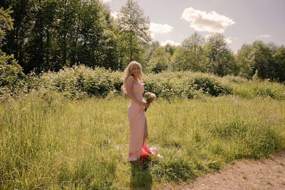 Blonde woman in pink gown holding bouquet stands in lush green meadow on sunny day.
