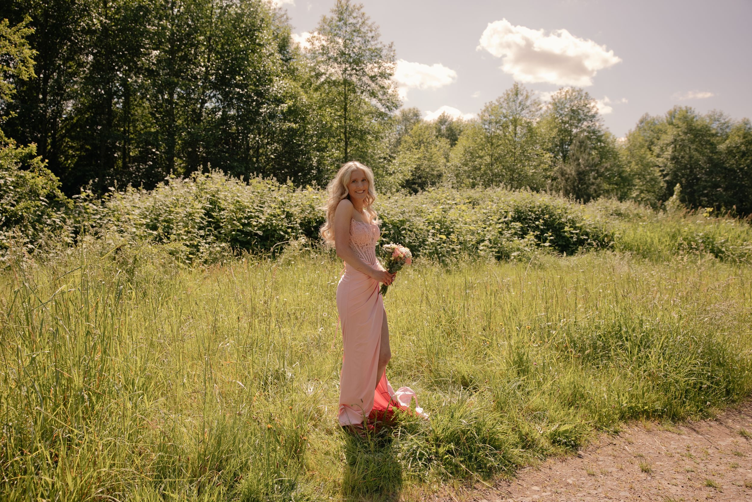 Blonde woman in pink gown holding bouquet stands in lush green meadow on sunny day.