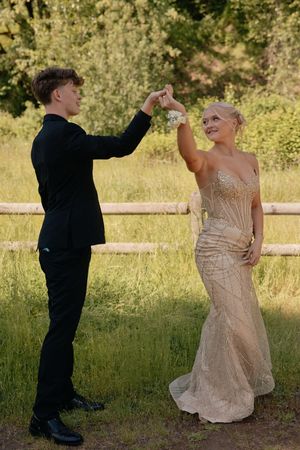 Young couple in formal prom wear dancing outdoors, man in black suit spinning woman in gold sequin gown.