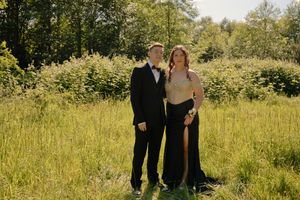 A couple in formal attire poses in a sunny green meadow, man in tuxedo and woman in elegant black gown.