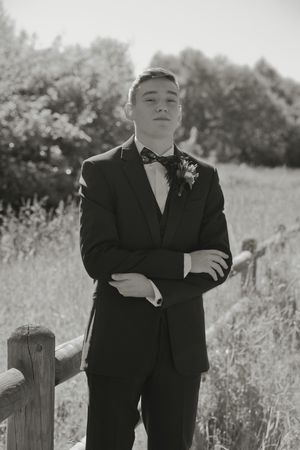 Young man in black suit with bow tie and boutonniere poses arms crossed by rustic fence in black and white photo.