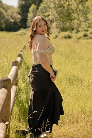 Young woman in black skirt and off-shoulder top posing by rustic wooden fence in sunny green meadow.