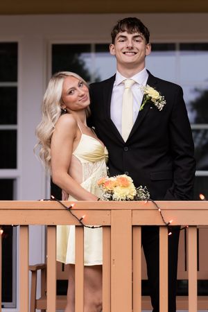 A couple in formal attire poses together on a wooden deck railing during what appears to be a prom or formal event.