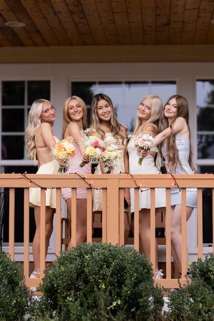 A group of bridesmaids in light colored dresses pose together on a wooden deck holding colorful floral bouquets.