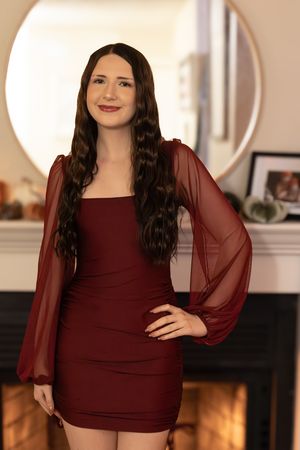 A figure in a burgundy bodycon dress with sheer long sleeves poses in front of a fireplace and round mirror.