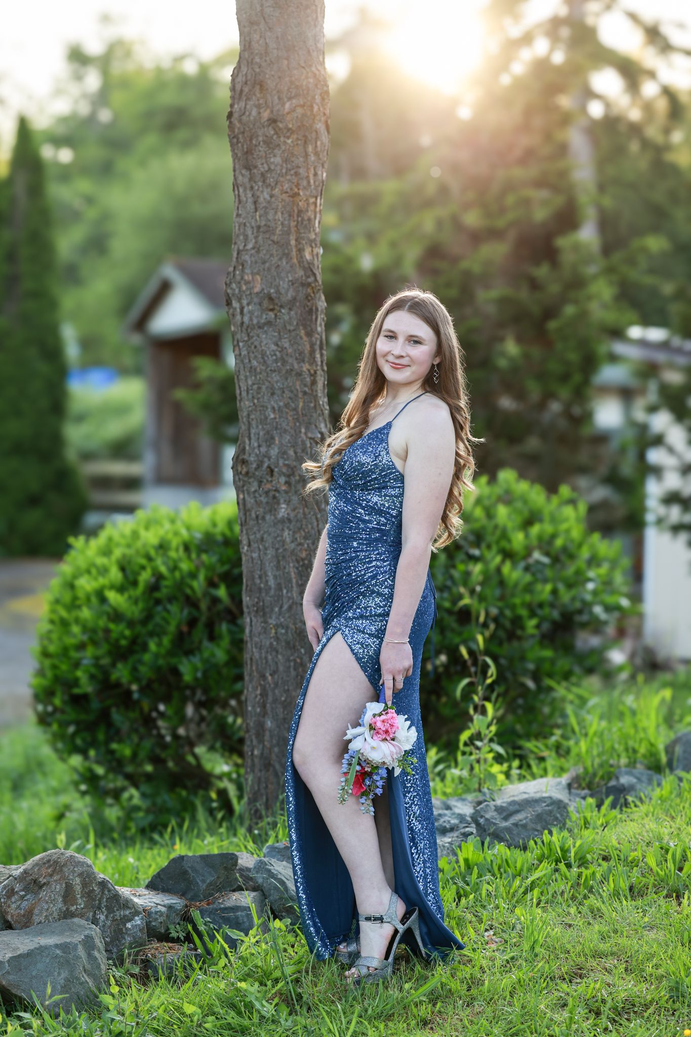A woman in a blue dress poses outdoors at sunset among trees and greenery for a portrait session.