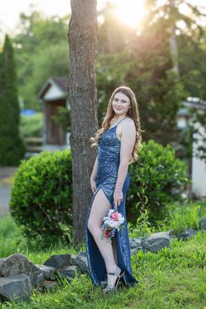 A woman in a blue dress poses outdoors at sunset among trees and greenery for a portrait session.