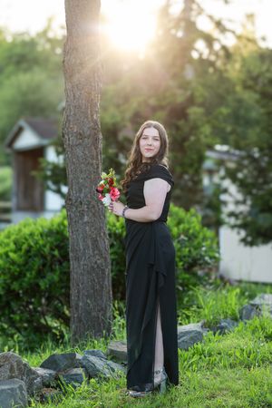 A person in a long black dress poses thoughtfully against a tree trunk at sunset in a garden setting.