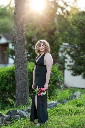 A person in a black dress poses on a grassy hill during golden hour with trees and sunlight in the background.