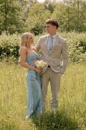Couple in formal attire standing in a green meadow, woman in blue dress holding bouquet, man in beige suit.