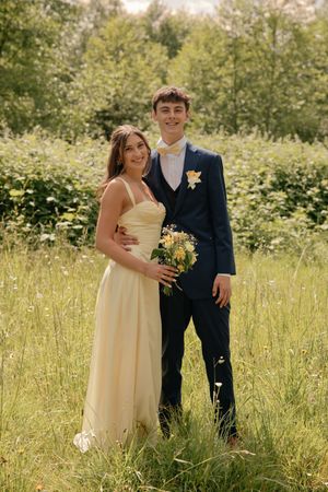 Young couple in prom attire posing outdoors, woman in yellow gown holding bouquet, man in navy suit with boutonniere.