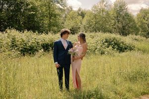 Young couple dressed for prom standing in a lush green field, woman holds a floral bouquet.