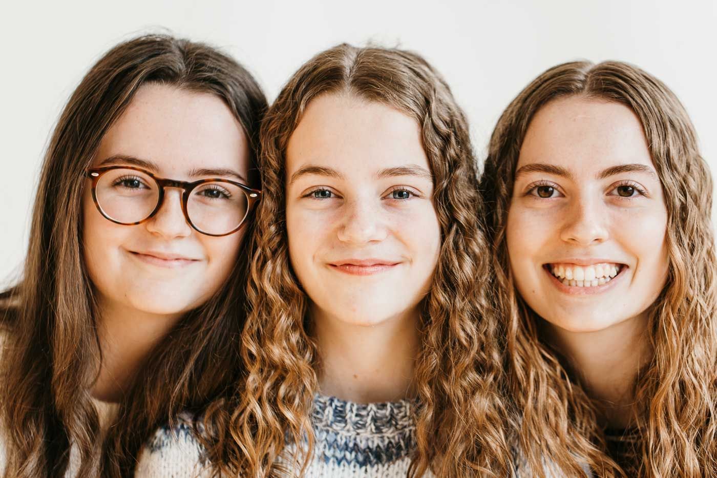 Three young people with wavy brown hair smile warmly at the camera, with one wearing glasses and a casual gray sweater.