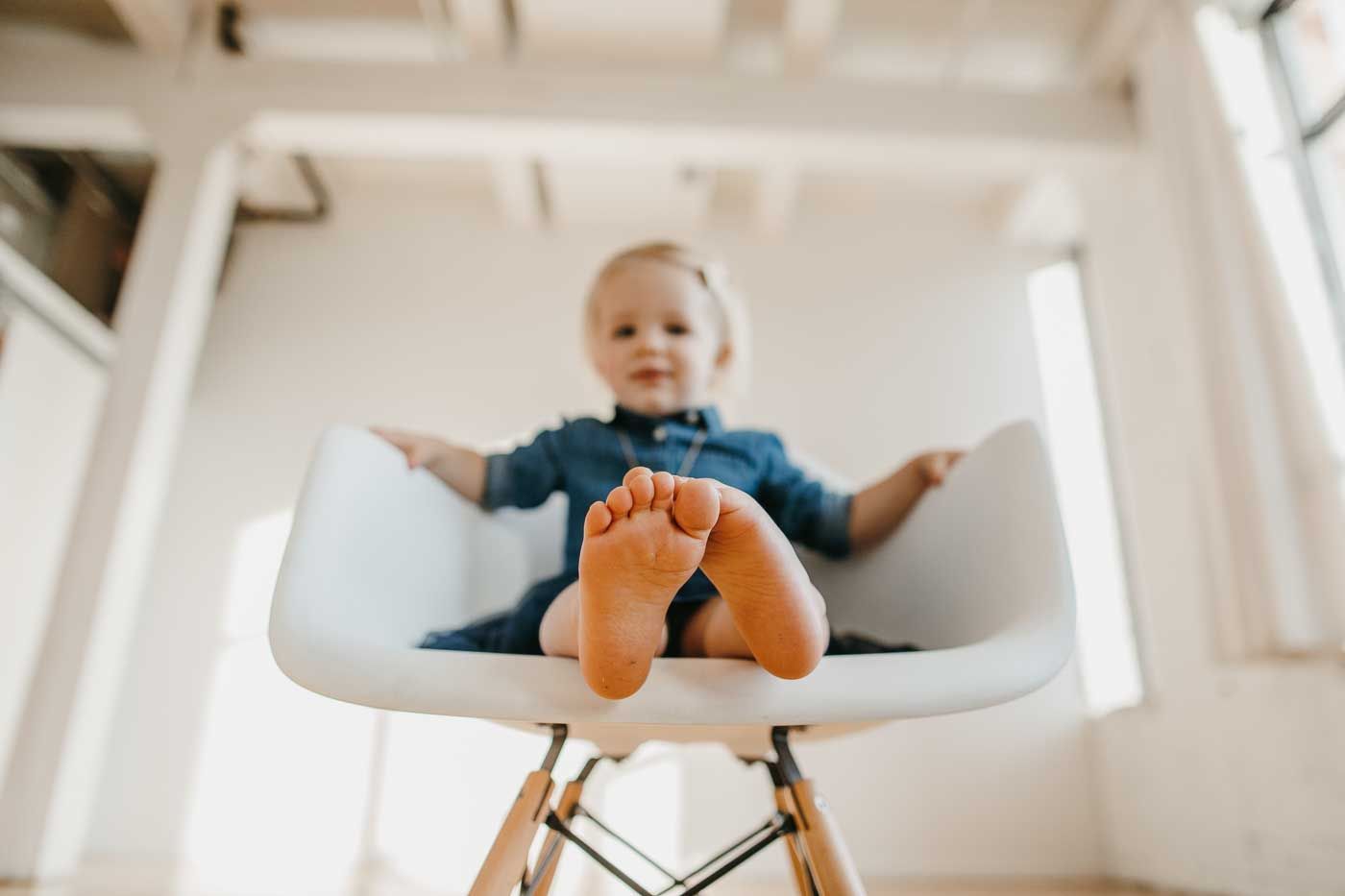 Looking up from below at a child sitting in a white Eames-style chair with bare feet against a bright interior.