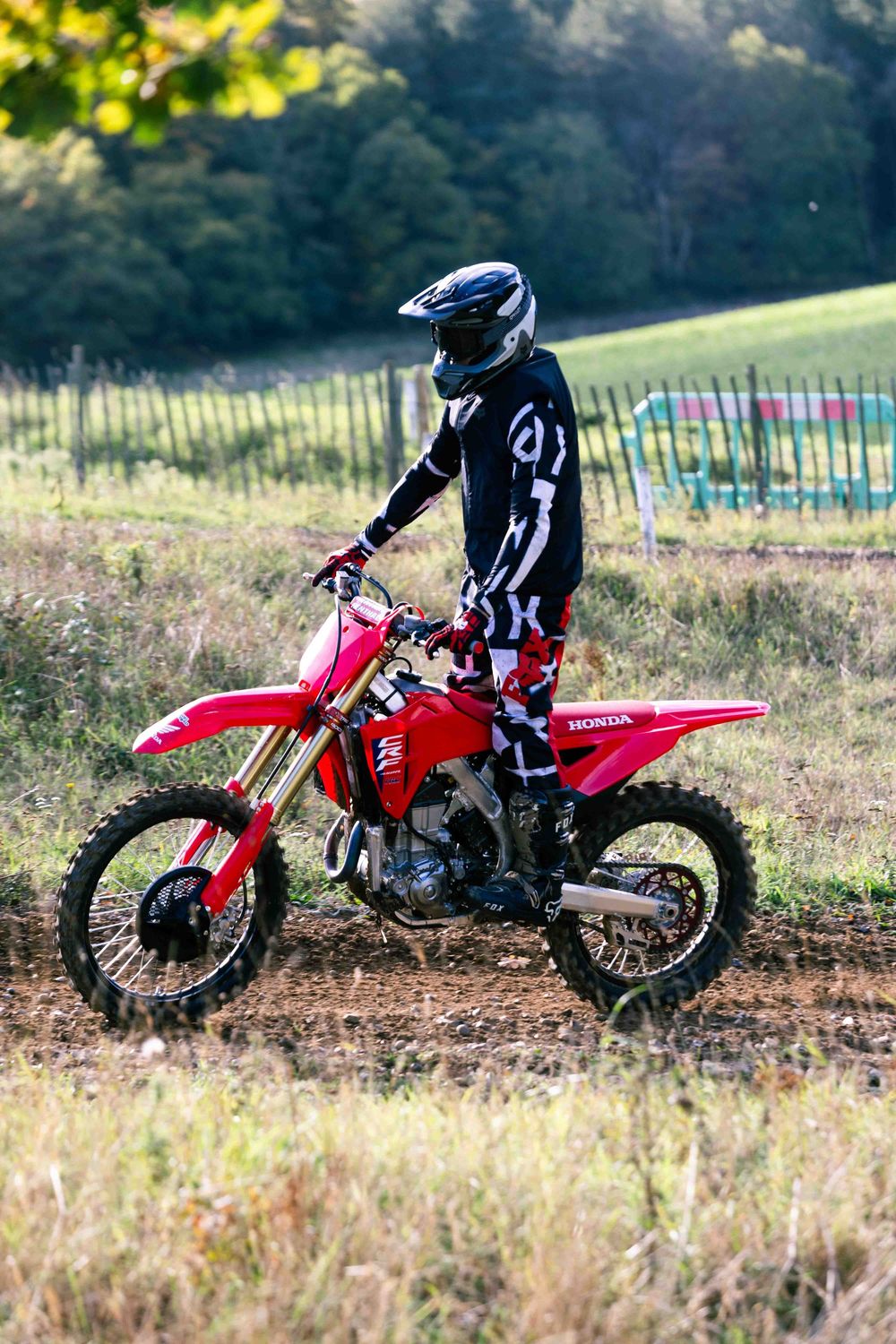 A dirt bike rider in black protective gear stands with their red Honda motorcycle on a grassy hill during daytime.