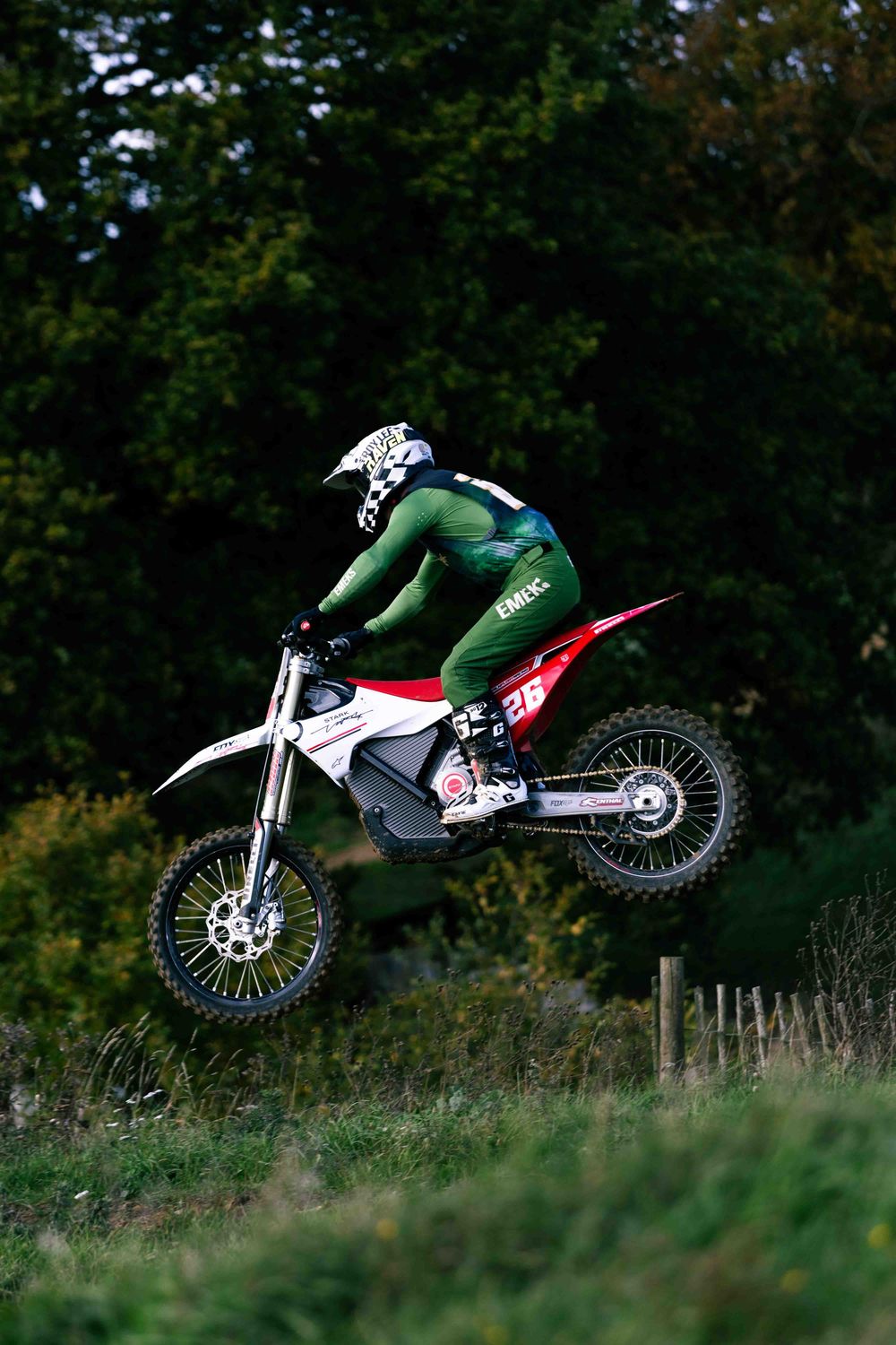 Sequence of a dirt bike performing a dramatic jump over a grassy hill against a dark forest backdrop.