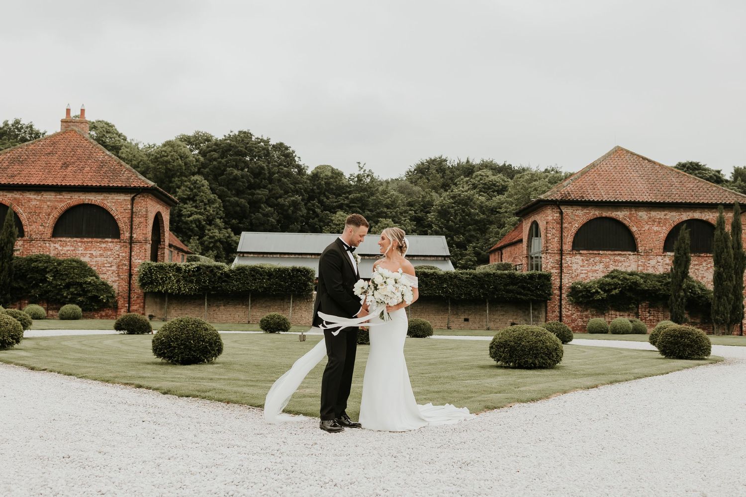 A couple in wedding attire playfully walks and dances together on a gravel path near a rustic brick building.