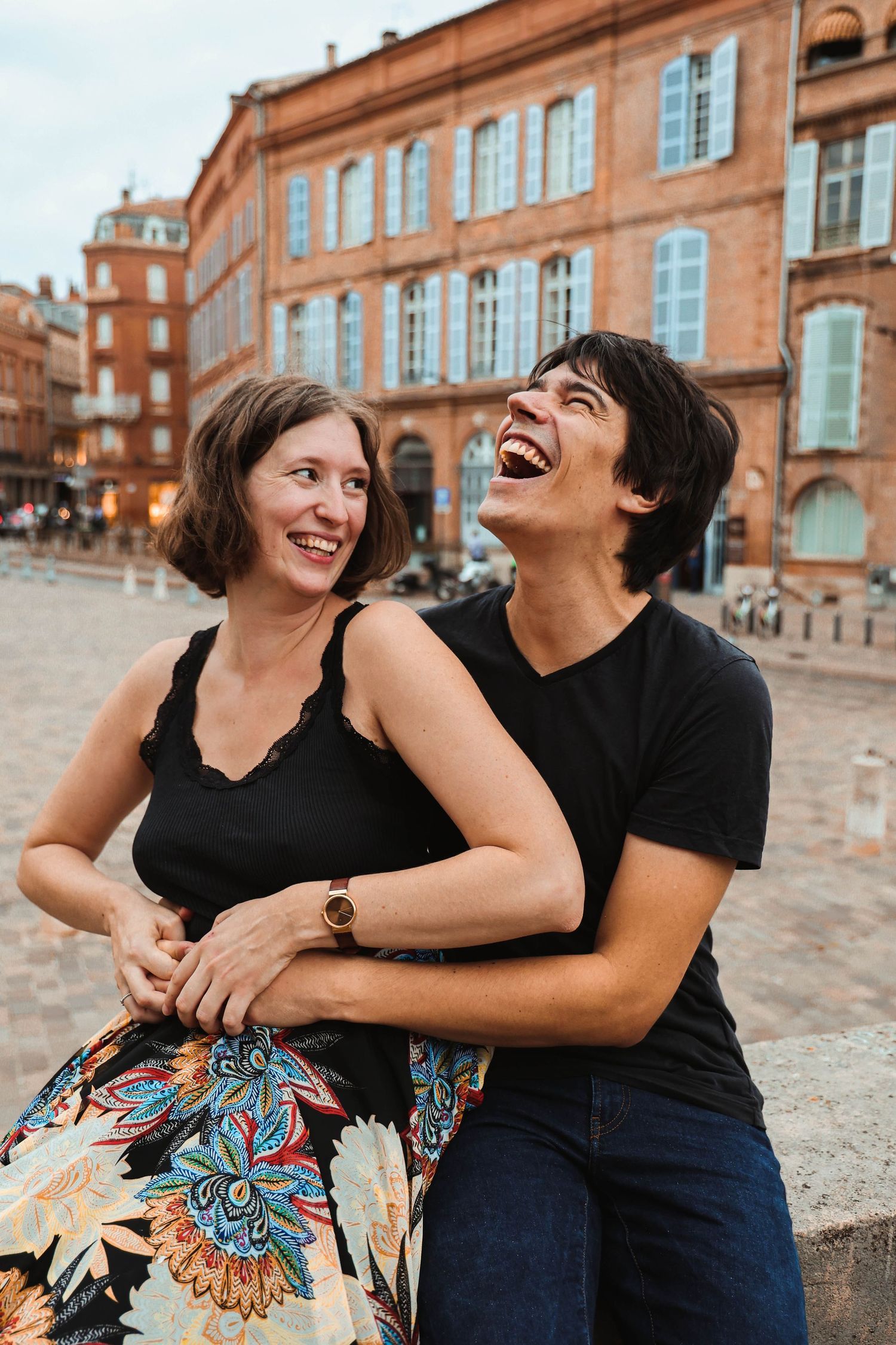 Joyful couple laughing together outdoors near historic brick building, woman in floral skirt embraced by man.