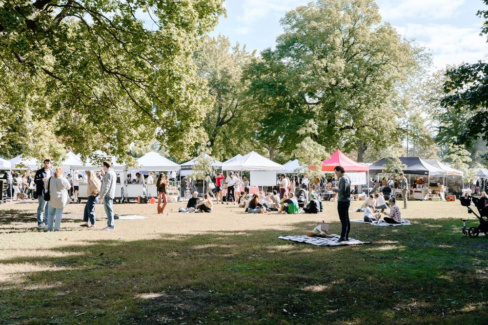 People enjoying a sunny day in a park with scattered tents and canopies set up under large shady trees.