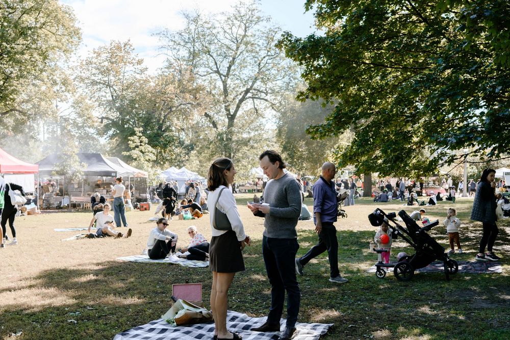 A sunny outdoor gathering with picnic blankets spread under trees in a park setting.