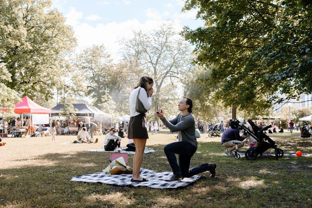 A couple shares a special moment on a picnic blanket in a sunny park with festival crowds in the background.