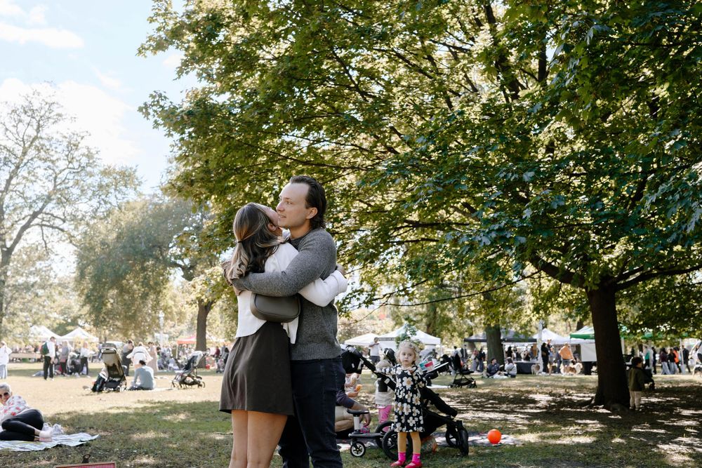 A couple embraces in a sunny park on a fall day with trees and parked bikes in the background.