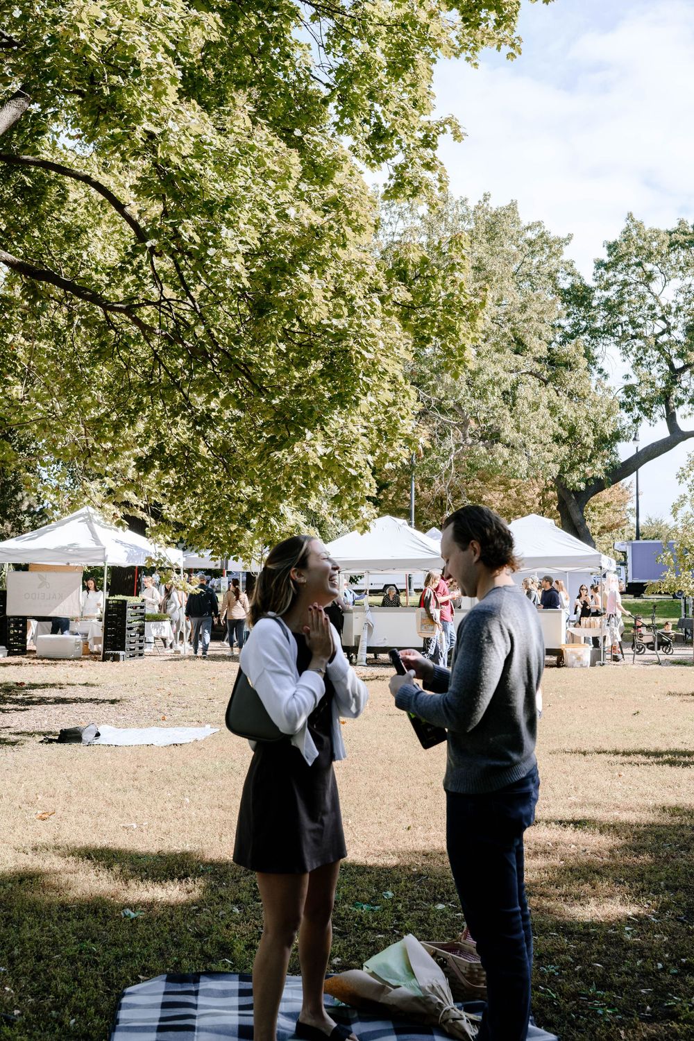 Candid outdoor scene in a park with people interacting under tall trees and white tents in the background.