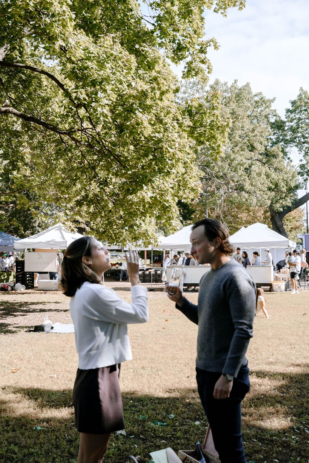 A couple playfully interacts on a grassy field with white tents visible in the background on a sunny autumn day.