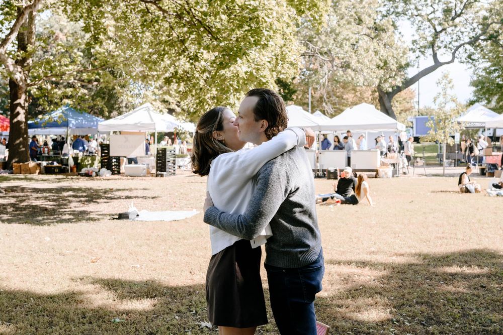 People gather at an outdoor event with white tents and trees visible in a park-like setting.