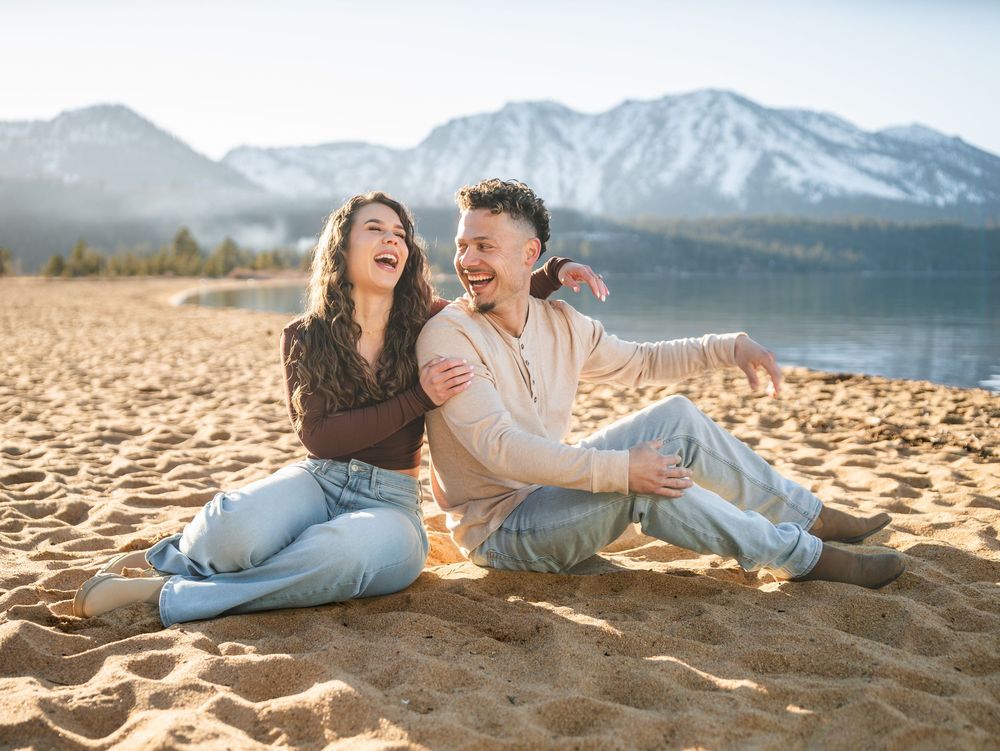 A couple relaxes and laughs together on a sandy beach with snow-capped mountains in the background.