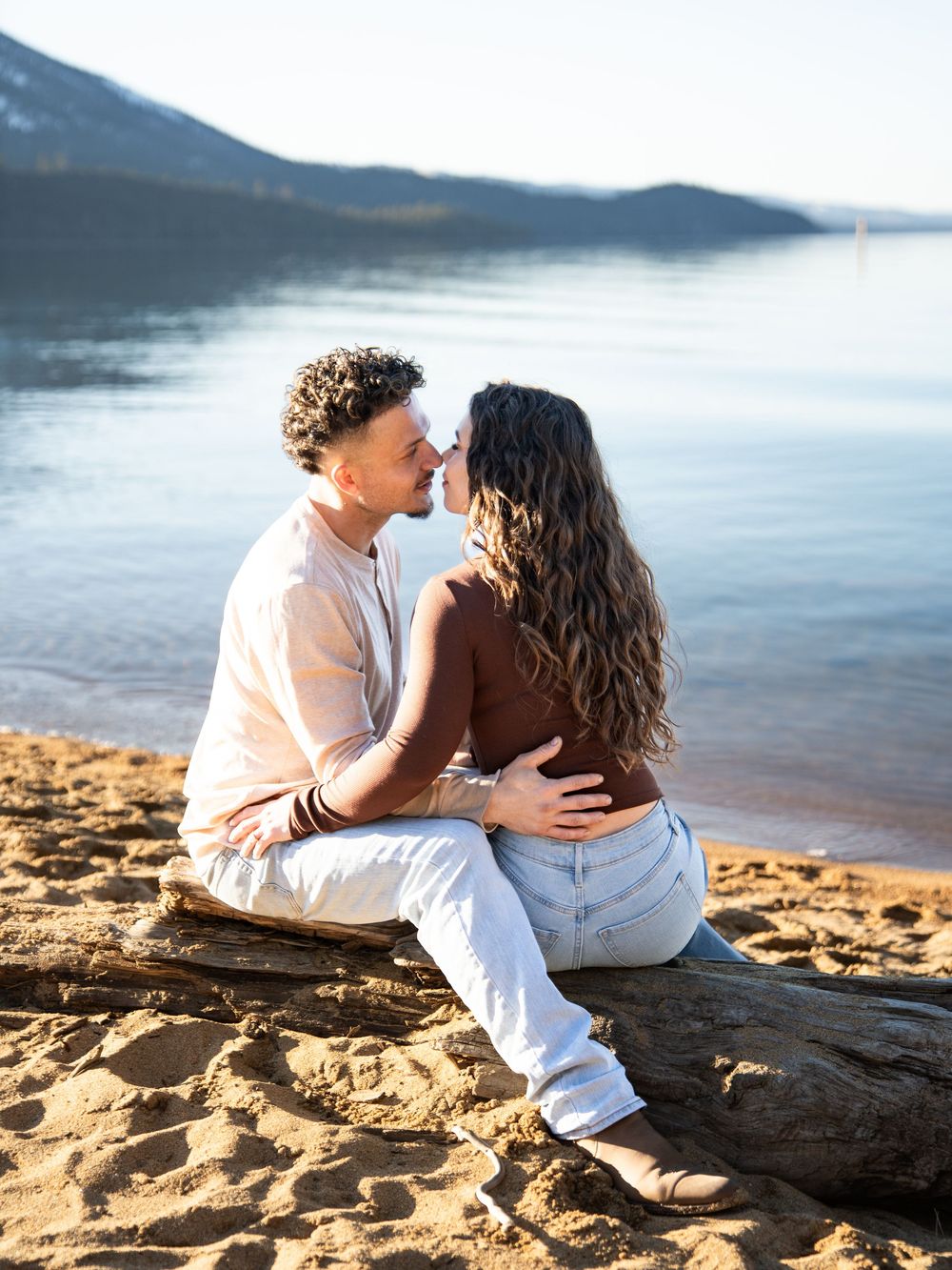Two people share an intimate moment while sitting on driftwood at a lakeside beach during golden hour.